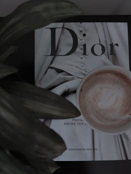 Flat lay of a cappuccino next to Dior book, creating a stylish and cozy ambiance.