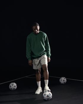 A man in a green sweatshirt practices soccer skills indoors with multiple soccer balls.