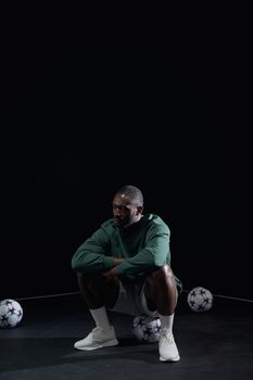 A focused African American man in sports attire squats in a studio setting with soccer balls around him.