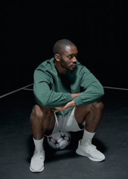 African American man in sportswear sits on a soccer ball on an indoor court.
