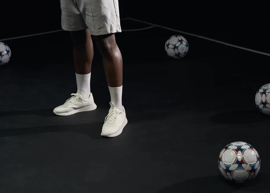 Joueur en short gris entouré de ballons de foot sur terrain sombre minimaliste