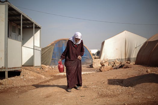 An elderly woman walking in a Syrian refugee camp, carrying a red container.