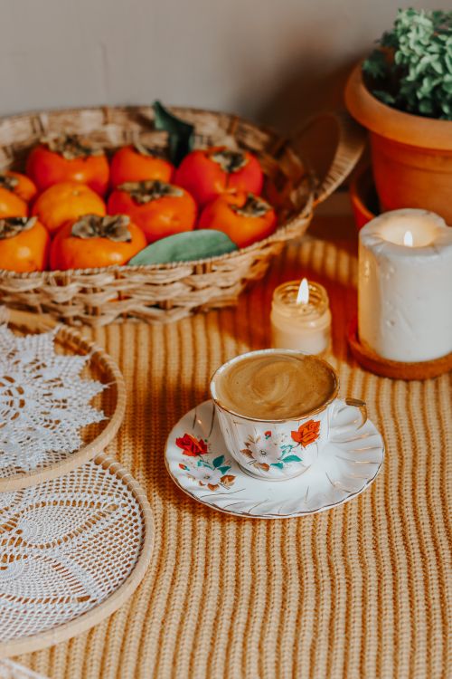 Free Warm, inviting autumn scene featuring cappuccino, persimmons, and candles on a woven tablecloth. Stock Photo