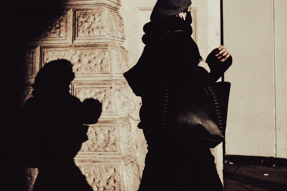 Stylish woman in a beret and overcoat walking with a distinct shadow against a textured wall.