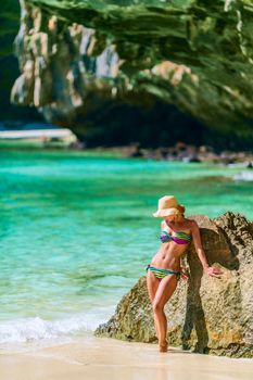 Woman in a colorful bikini and sun hat posing on a tropical beach with turquoise water.