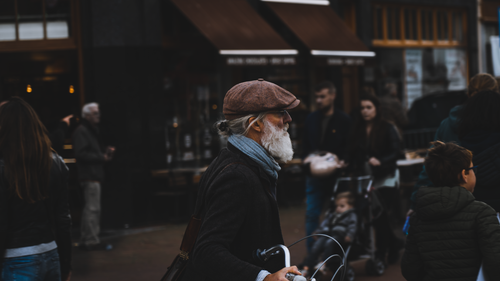 Free Elderly man with a white beard, wearing a brown hat, navigating a busy city street with a bicycle. Stock Photo