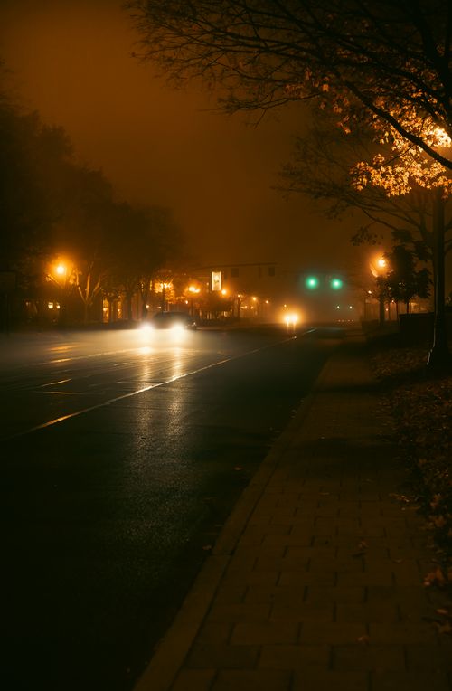 Free Capture of a foggy night with streetlights illuminating a quiet road, creating a moody atmosphere. Stock Photo
