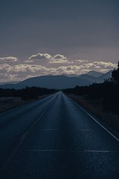 A calm road leading towards distant mountains under a cloudy dusk sky.
