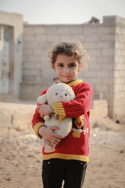 Free A young Middle Eastern girl holding a plush toy, standing outdoors in a Syrian town. Stock Photo