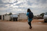 A girl with a backpack walks on an unpaved road in Idlib, Syria, under a cloudy sky.