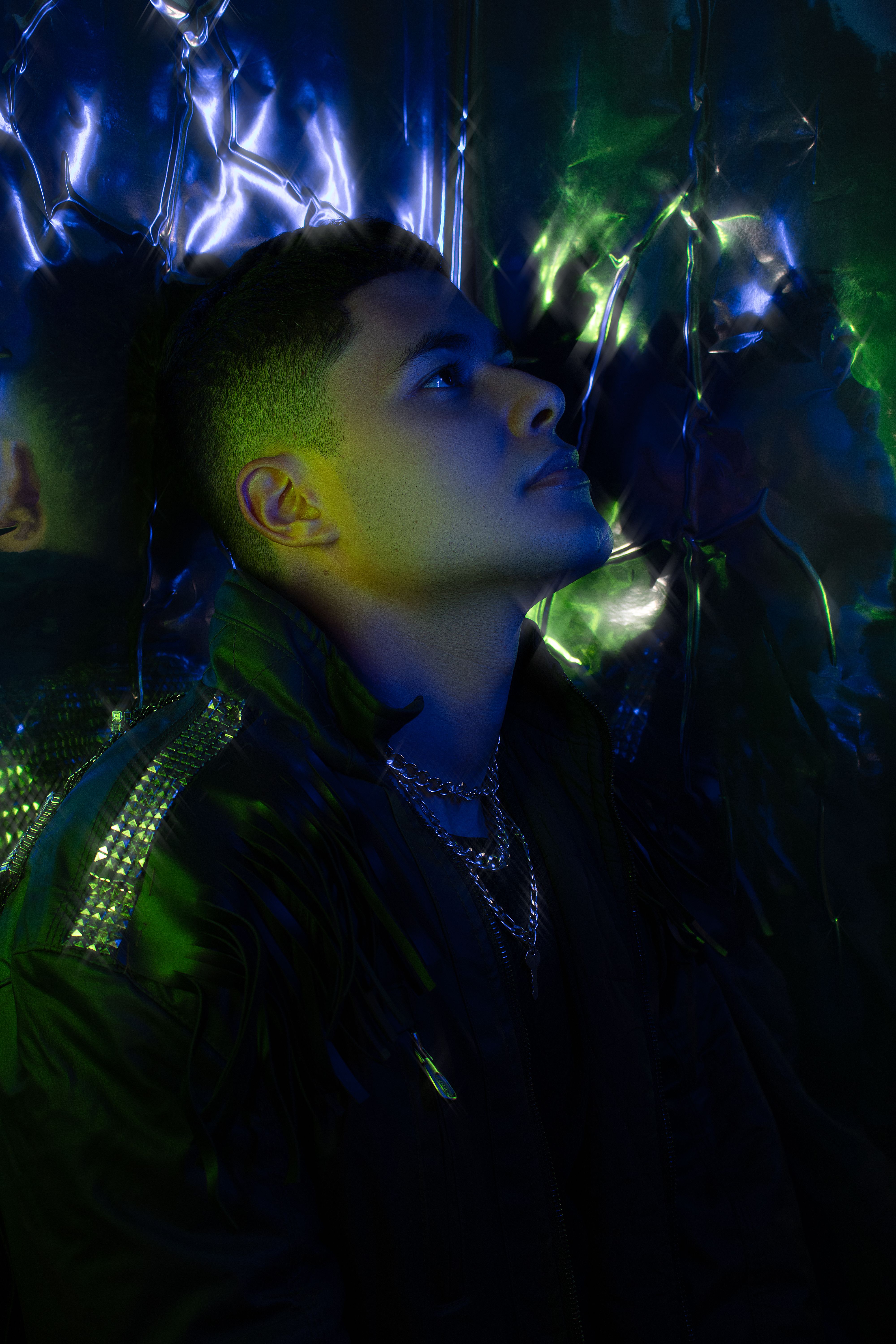 A striking portrait of a man in vibrant blue and green lighting, wearing a necklace.