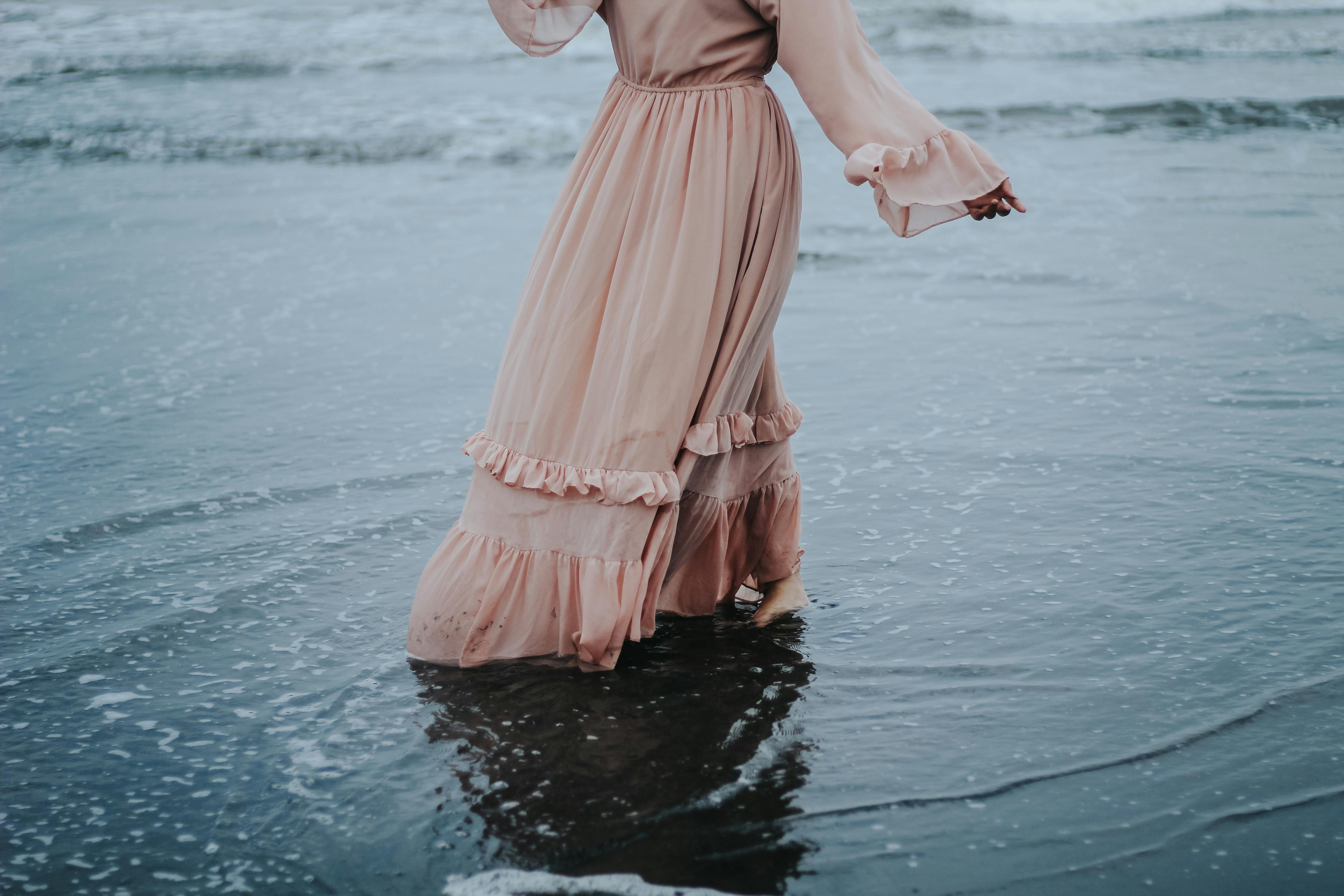 A woman in a flowing dress walks serenely through ocean waves at the beach, capturing a tranquil moment.