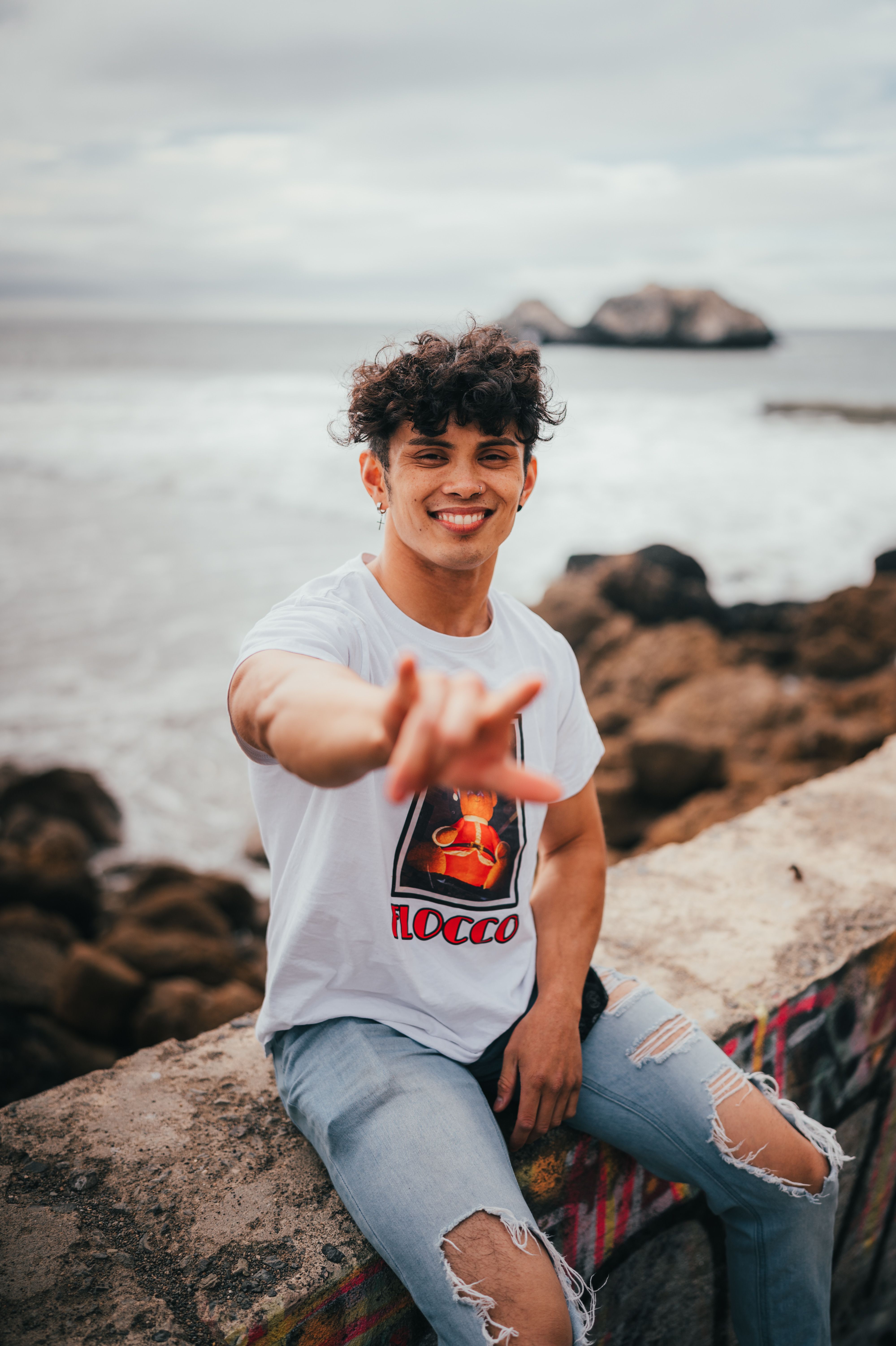 Smiling young man in a white shirt sitting by the ocean, making a hand gesture.
