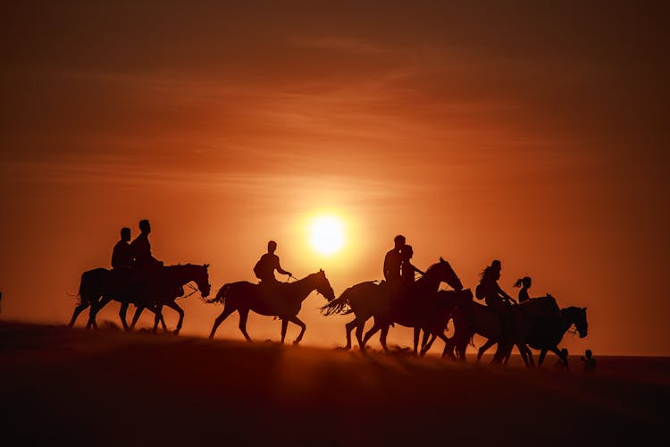 A Silhouettes Of Group Of People Riding Horses Over A Sunset