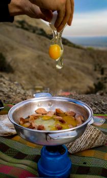 Hand cracking egg into a camp stove pan with sausages outdoors.