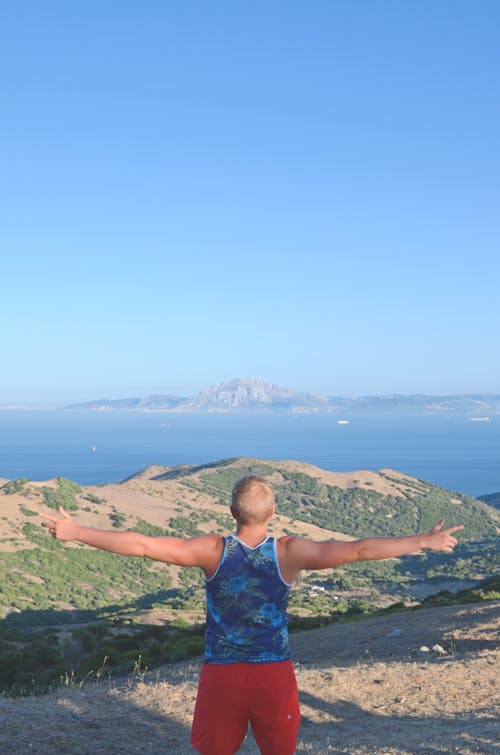 Uomo Che Indossa Canotta Blu Verde E Bianca In Piedi Sulla Montagna Marrone Vicino Al Corpo D'acqua Blu Sotto Il Cielo Blu Durante Il Giorno