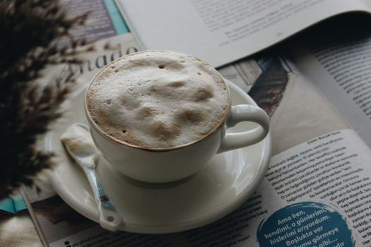 A steaming cup of foamy coffee resting on a saucer atop a spread of newspapers.