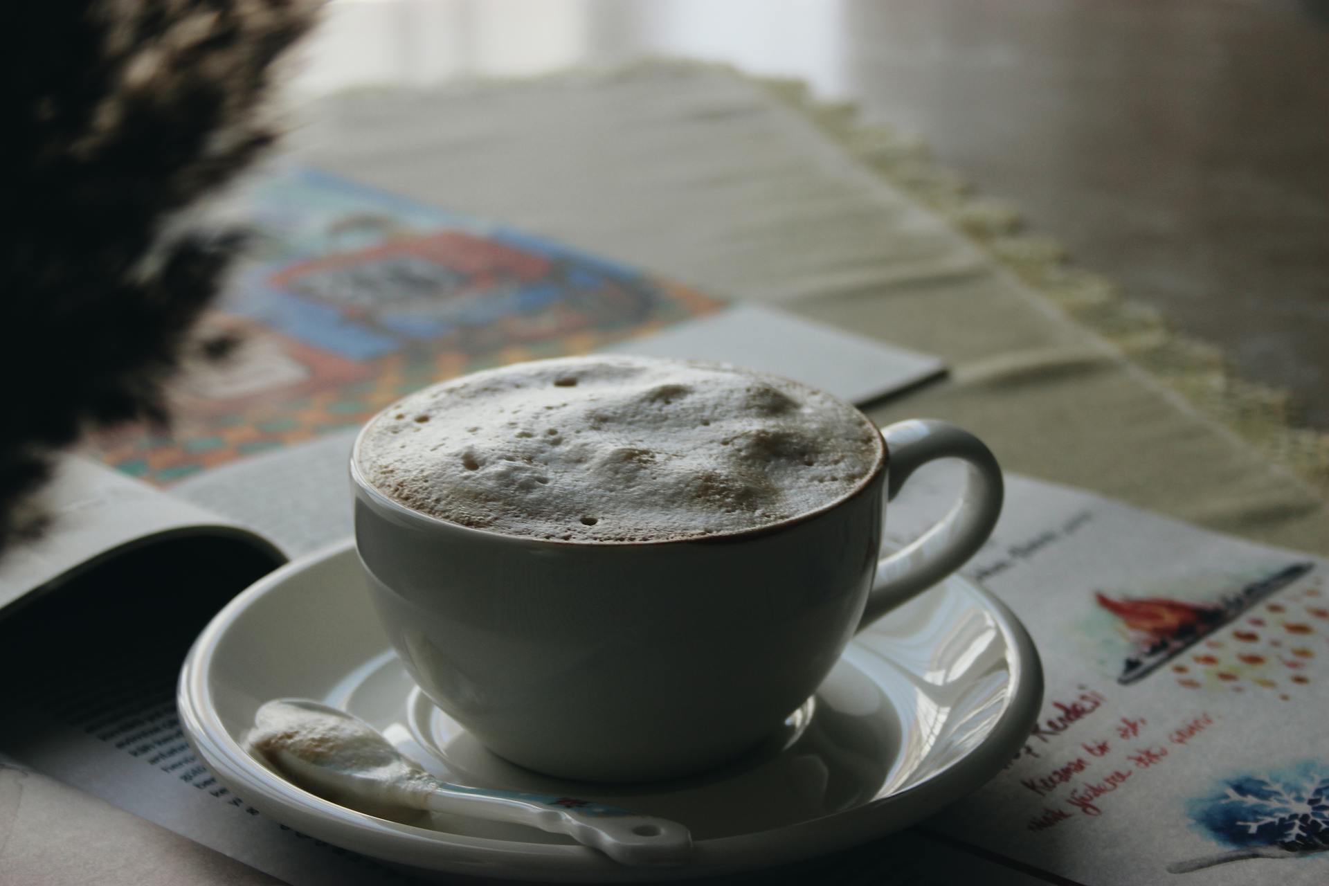 A warm cappuccino with foam in a cup on a saucer, set on a table with a magazine, creating a cozy cafe atmosphere.