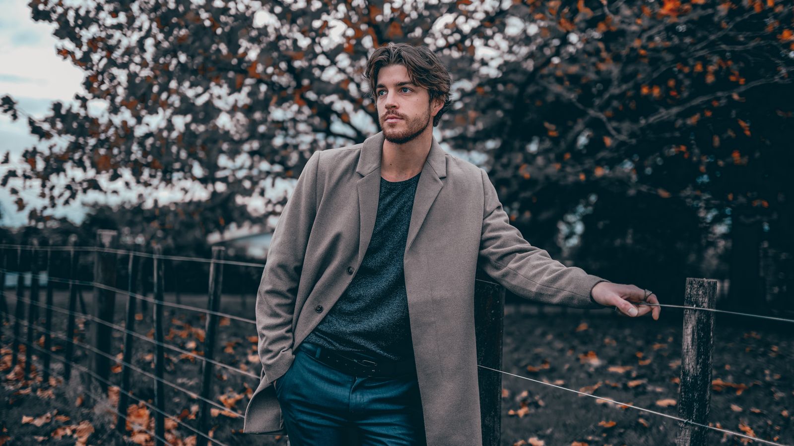 Man in a gray coat leaning on a fence in a fall setting with vibrant leaves