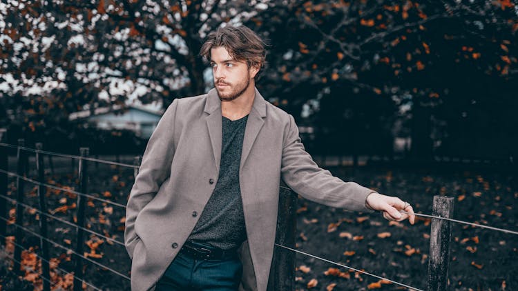 A Man In Gray Coat Leaning On A Wired Fence