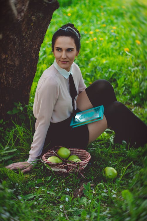 無料 茶色の大きな木の下で緑の草に座っている白いドレスシャツをしている女性 写真素材