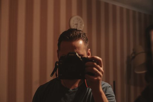 A man holding a camera, capturing his reflection in a dimly lit room with striped wallpaper.