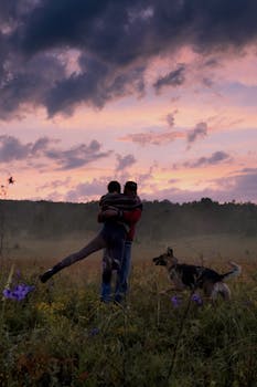 A couple embraces lovingly with their dog in a meadow at sunset, creating a warm silhouette.