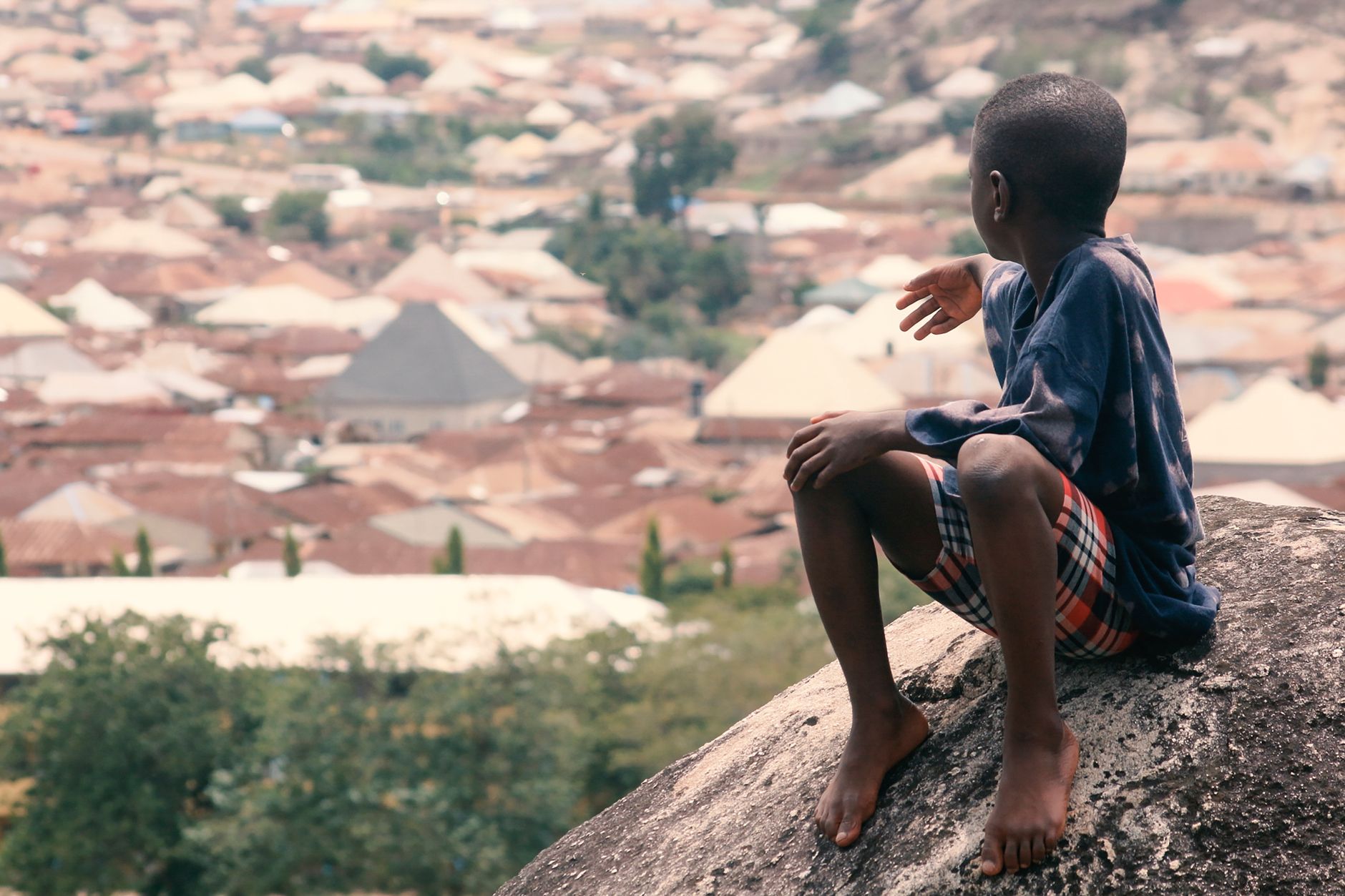 Young boy sits on a rock, overlooking a sprawling cityscape. Serene moment captured.