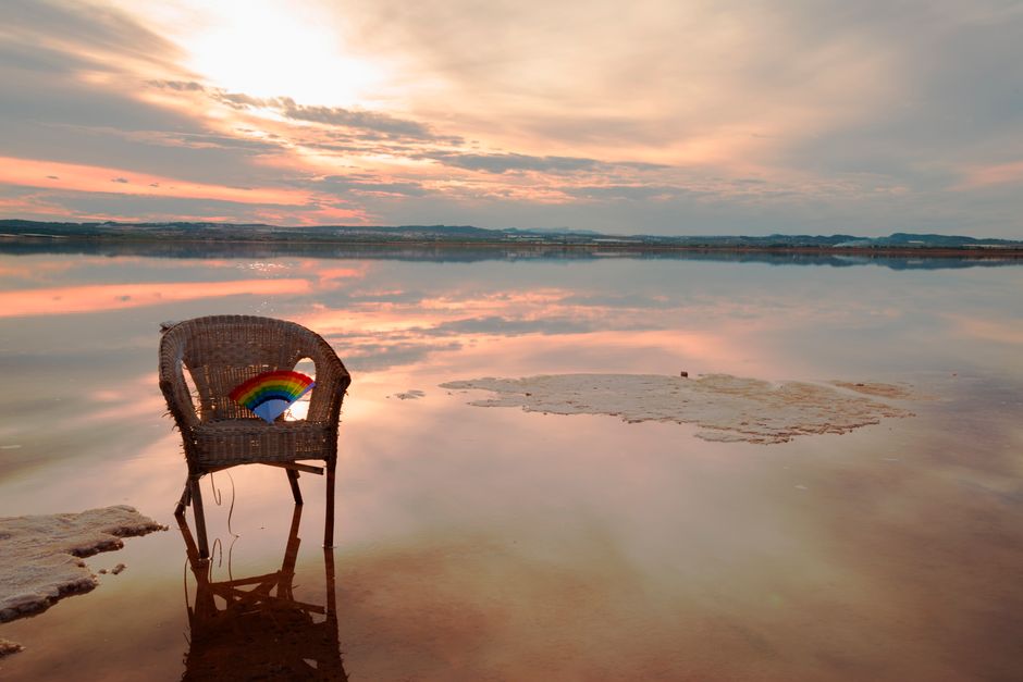 Woven chair by Laguna de Torrevieja at sunset, with serene water reflections and a rainbow hand fan.