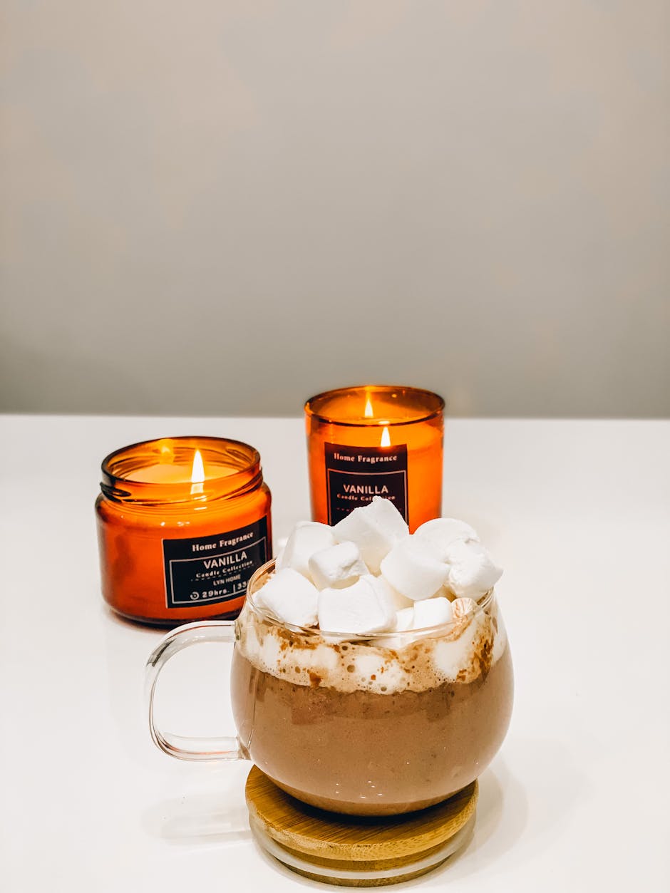 Warm hot chocolate topped with marshmallows beside lit vanilla candles on a white surface.