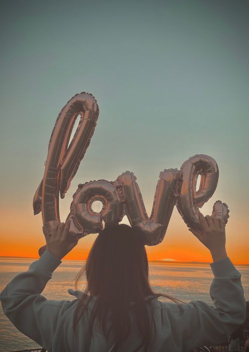 Free A woman holding love-shaped balloons at a beach during a stunning sunset creates a romantic atmosphere. Stock Photo
