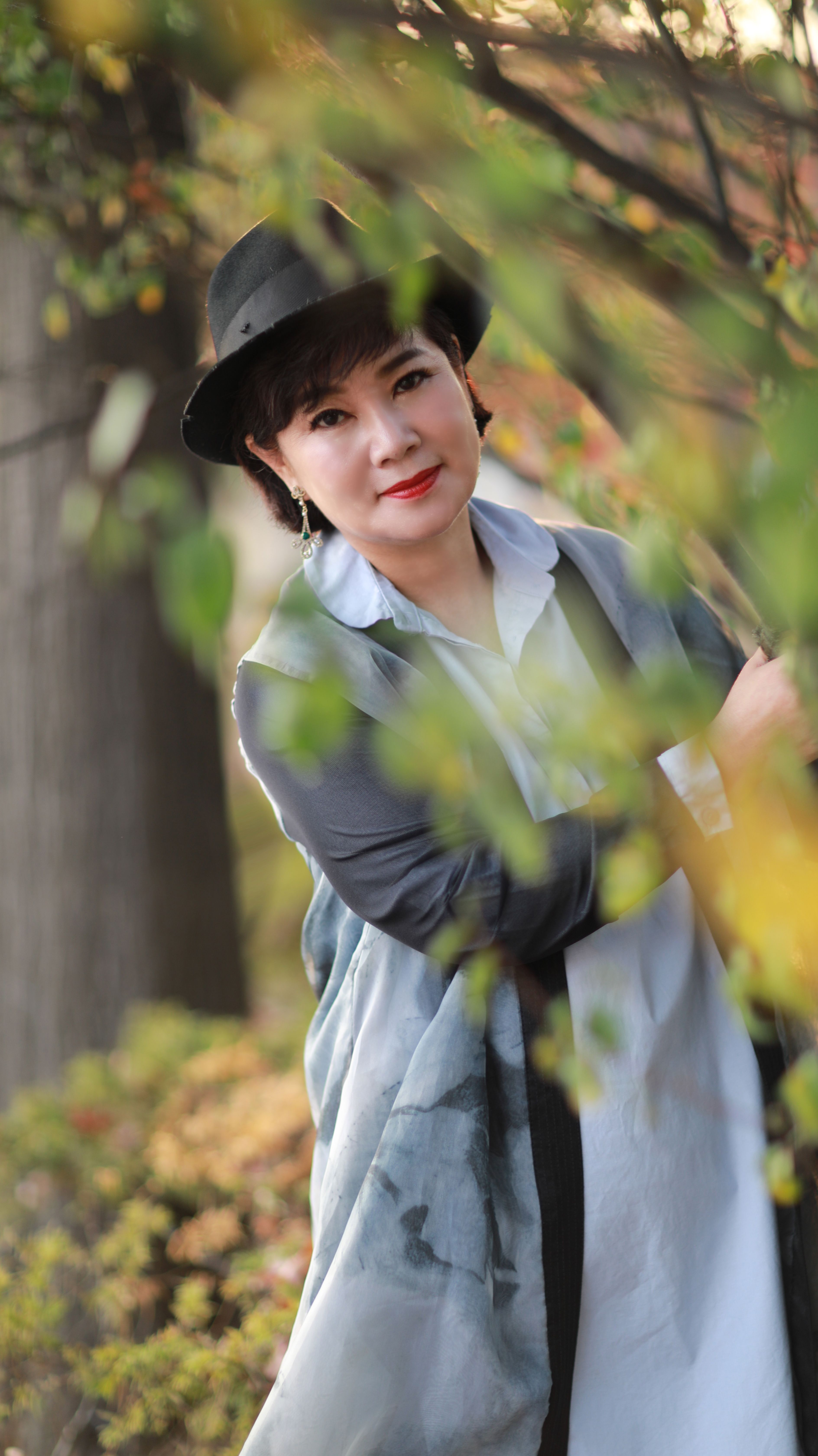 Chic woman smiling outdoors wearing a fedora and long sleeves amidst foliage.