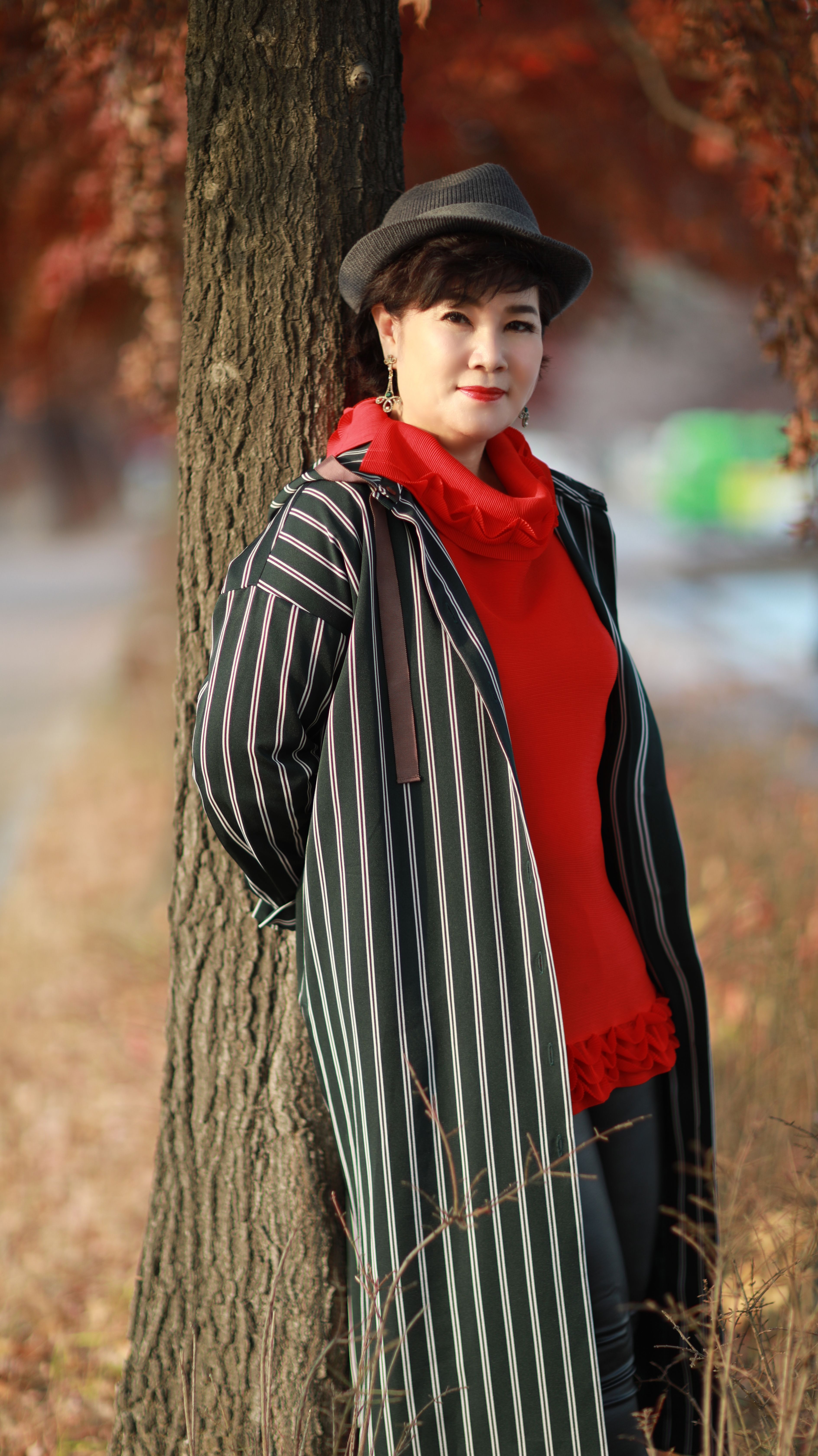 Woman in a fedora and striped coat standing by a tree in autumn.