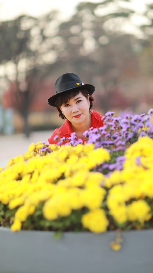 Woman in Red Shirt Wearing Black Fedora  Hat Standing Beside a Bunch of Purple and Yellow Flowers 