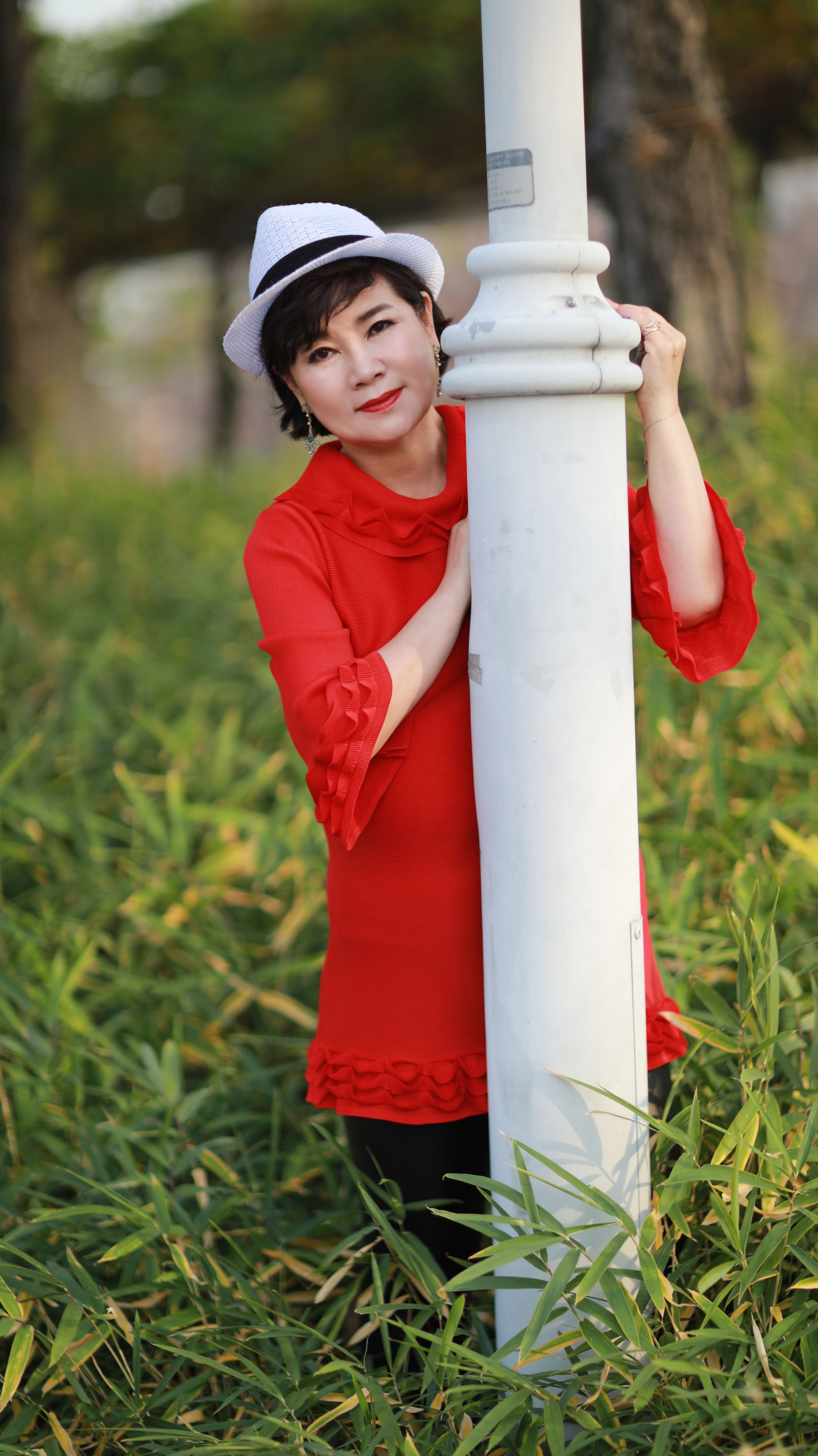 Woman in a red dress and fedora hat leaning against a post in a grassy area.