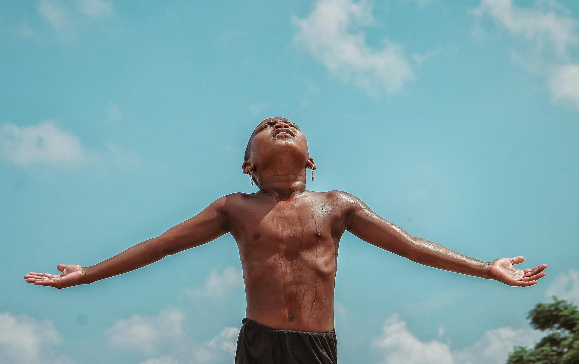 A shirtless boy enjoying the outdoors with arms wide open against a bright blue sky, expressing freedom.