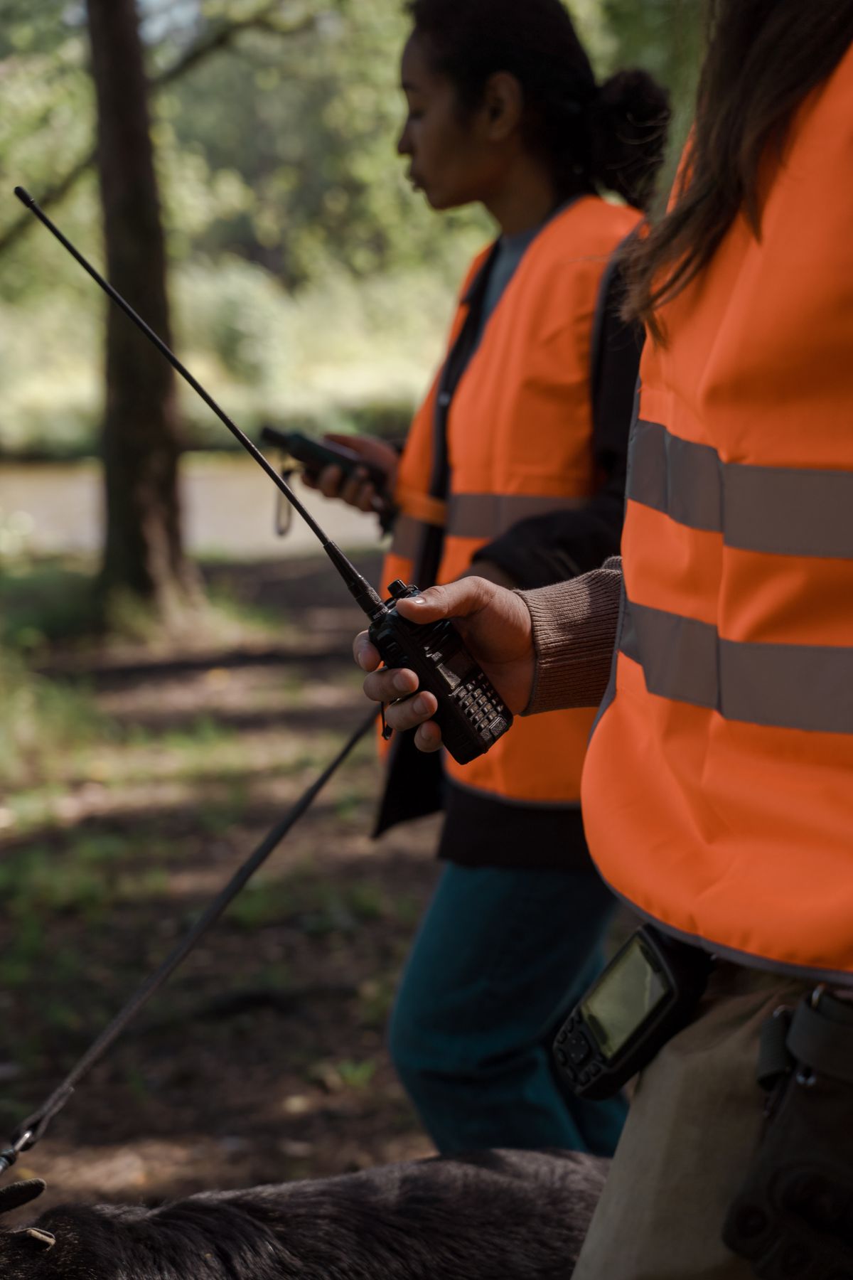 Hiker in an orange safety vest holding a handheld two-way radio with a long antenna on a wooded path