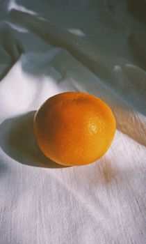 A single ripe orange placed on a white fabric with natural light and shadows creating a serene still life.