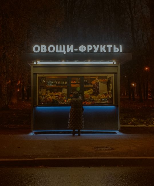 Illuminated kiosk with fresh produce at night, person standing in front, urban setting.