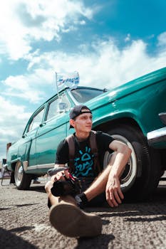 A young man sits next to a classic car under a blue sky, enjoying a sunny day.