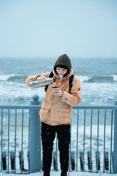 A young man pours hot tea from a thermos by the stormy sea, staying warm in winter clothing.