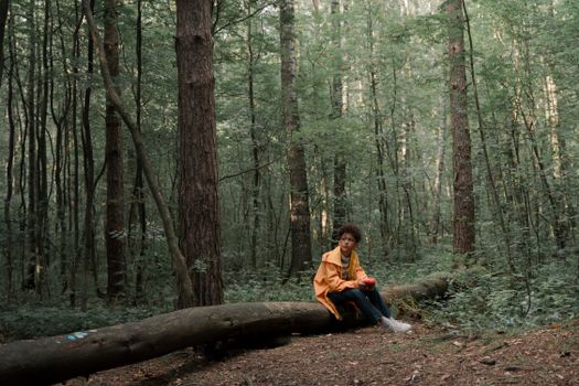 A young man wearing a yellow raincoat sits on a fallen log in a dense forest.
