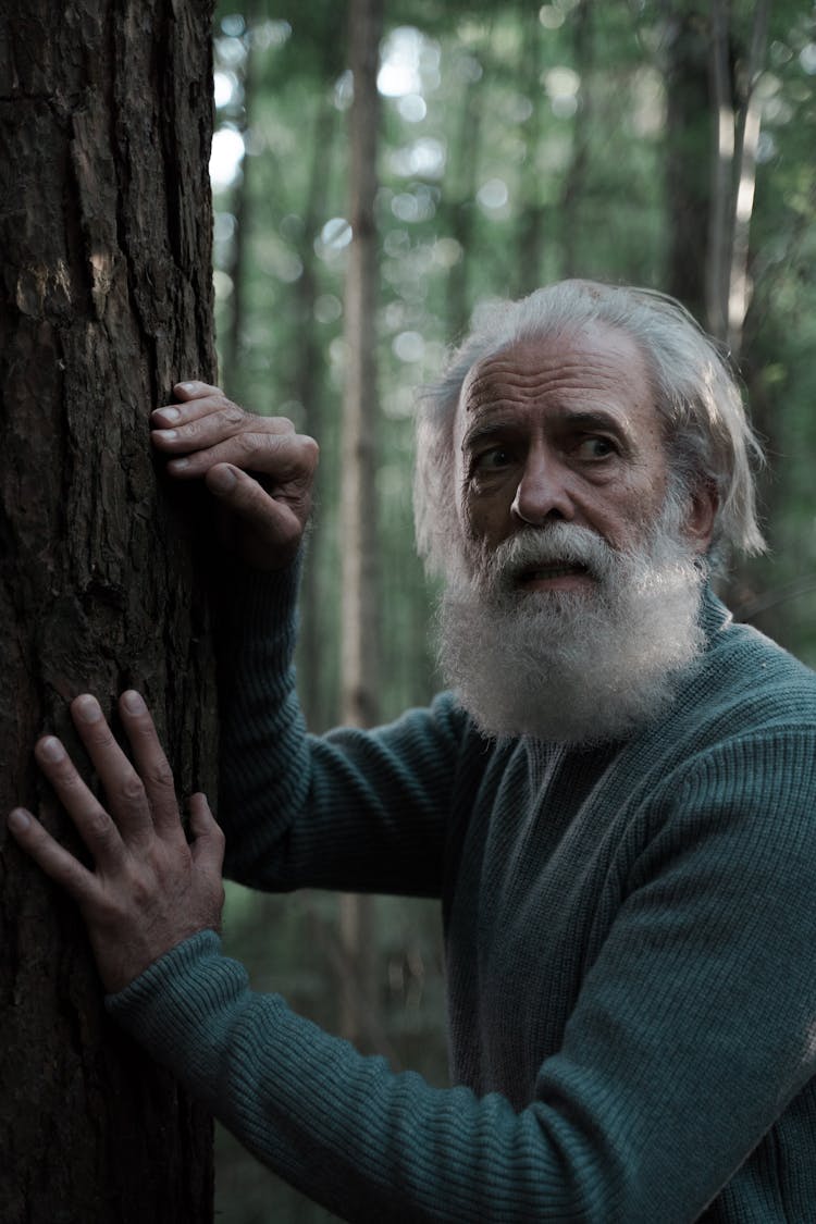 Bearded Man In Green Sweater Looking Scared While Touching Tree Bark