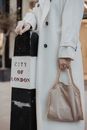 Woman in White Coat Holding Handbag Leaning on Concrete Post
