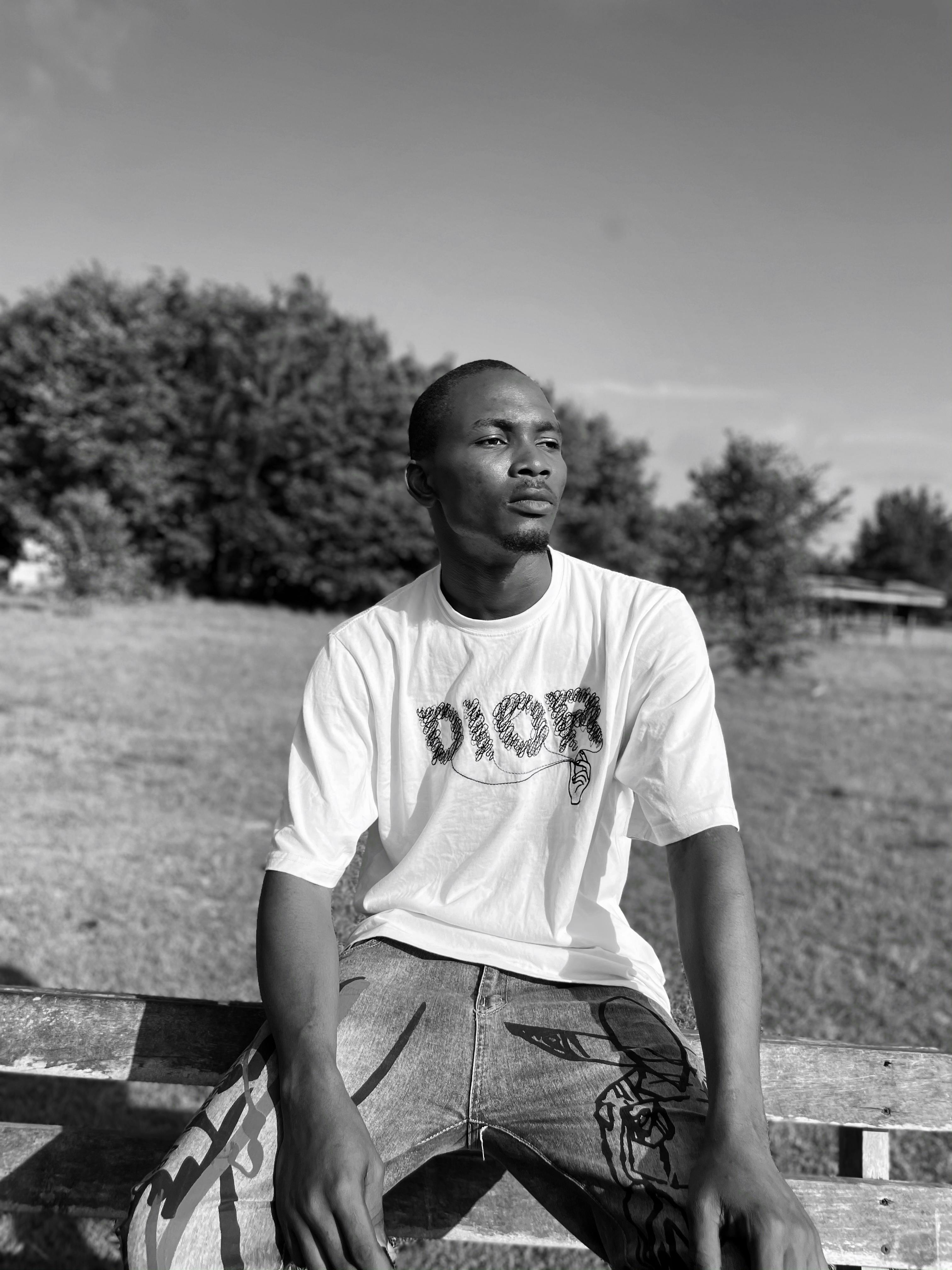 Grayscale photo of a man wearing a t-shirt, seated in an open field.