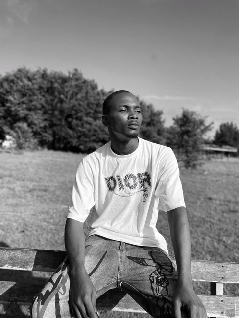 Grayscale photo of a man wearing a t-shirt, seated in an open field.