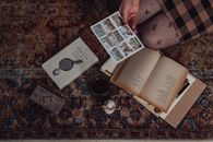 Hand Holding Postcard Above Books and Various Keepsakes Laying on Carpet