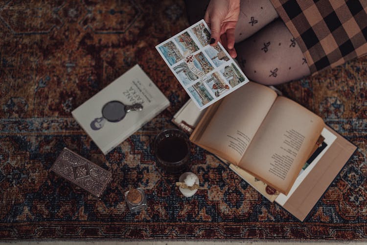 Hand Holding Postcard Above Books And Various Keepsakes Laying On Carpet