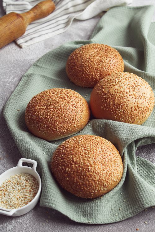 Free Close-up of four sesame seed buns displayed on a textured green cloth with a sprinkle of seeds nearby. Stock Photo