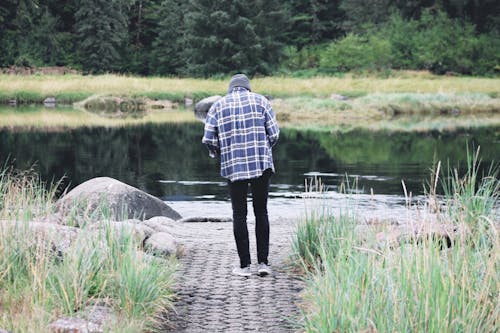 Back view of unrecognizable slim male in blue and white plaid shirt and skinny jeans standing on footpath near calm river with thick forest on other side
